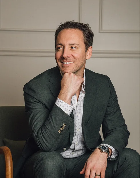 A candid portrait of Dr. Fournier sitting in a dark grey suit and a checkered button-down shirt. He is resting his chin on his hand and looking off-camera with a thoughtful smile.