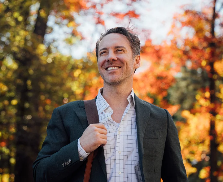 A medium shot of Dr. Fournier outdoors during autumn, wearing a green blazer and a checkered shirt with a brown leather bag strap over his shoulder. He is looking upward and smiling against a soft-focus background of vibrant orange and yellow fall leaves.