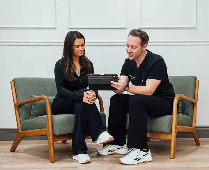 Dr. Fournier, wearing black scrubs, sits in a green armchair next to a female patient. He is holding a tablet and explaining information to her in a comfortable, professional lounge setting.