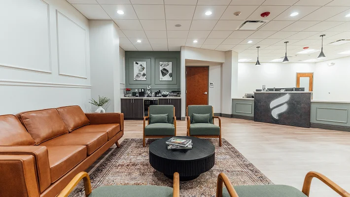 A wide view of the modern reception lounge at Fournier Plastic Surgery, featuring a tan leather sofa, green mid-century modern armchairs, and a black fluted coffee table. The background shows a coffee station and a sleek reception desk with a backlit logo.
