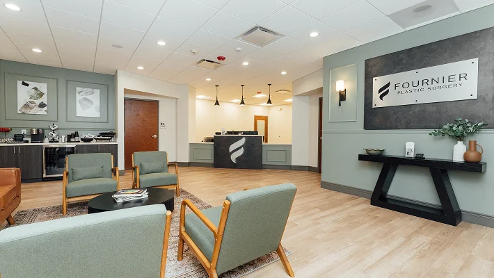 A view of the office lobby highlighting a large wall sign that reads "FOURNIER PLASTIC SURGERY" on a dark textured background. The room is decorated with sage green walls, elegant molding, and a minimalist black console table.