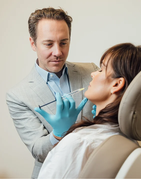 Dr. Fournier in a grey blazer and blue shirt, wearing blue nitrile gloves, carefully administering a facial injection to a patient reclining in a clinical chair.