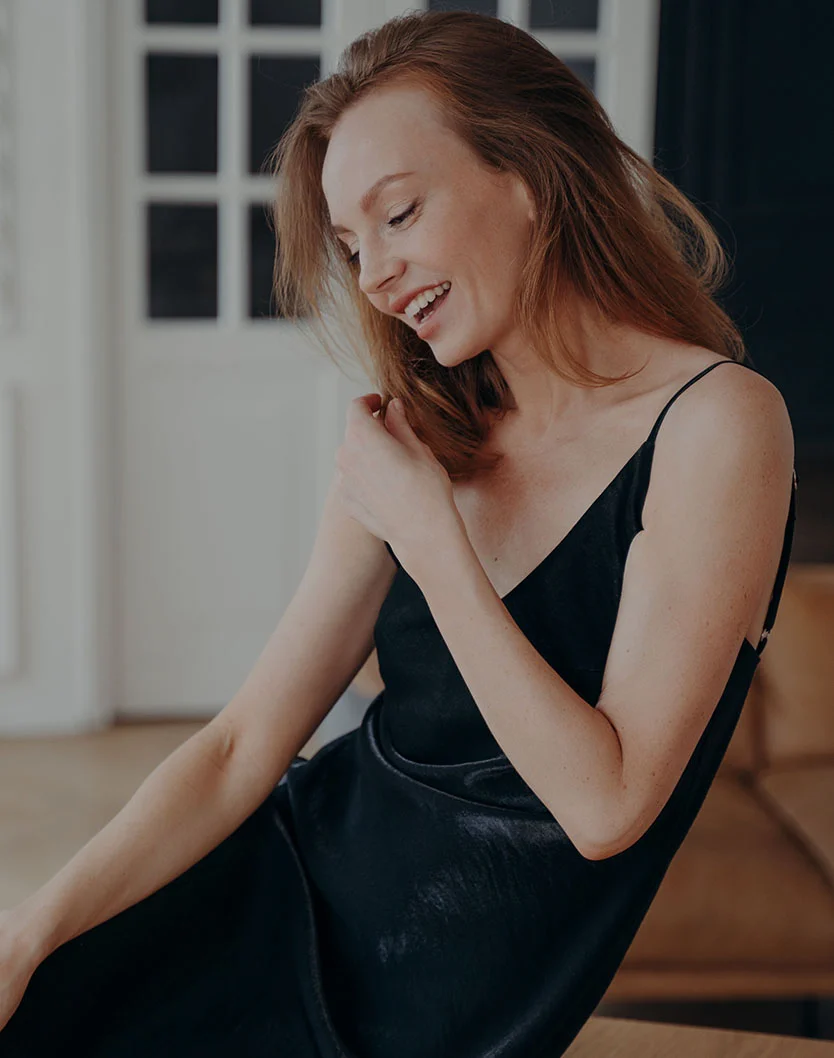A smiling woman with red hair wearing a black spaghetti-strap dress, looking down in a bright room.