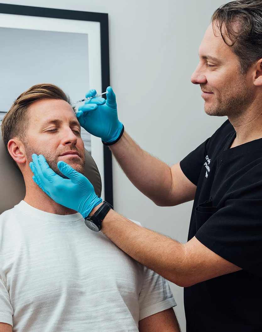 Dr. Fournier in a black shirt and blue gloves administering a facial injection to a male patient with his eyes closed.