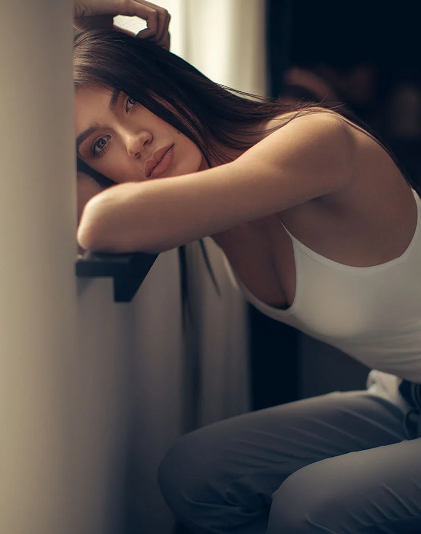 A woman with long dark hair in a white tank top leaning her arms on a ledge, looking thoughtfully into the distance.