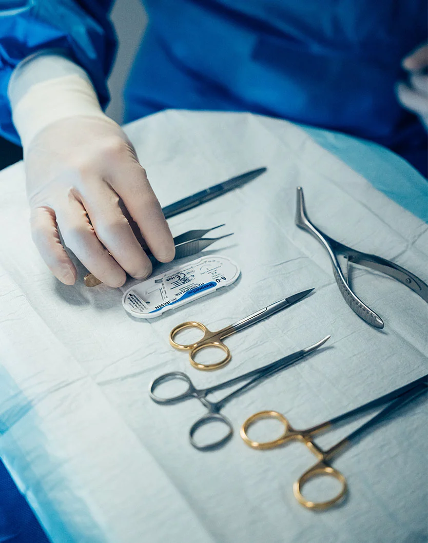 A close-up view of a sterile surgical tray containing precision medical instruments, including gold-handled scissors and forceps, as a surgeon in white gloves reaches for a tool.