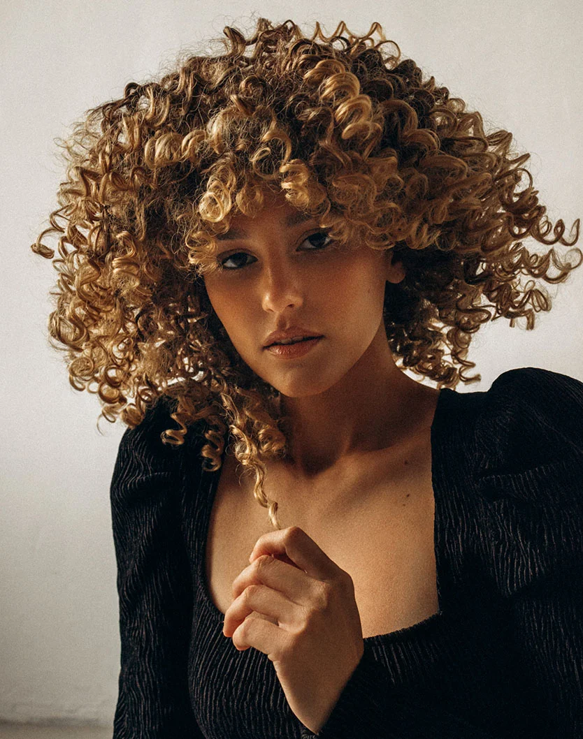 A woman with voluminous, curly blonde hair wearing a black textured top, looking toward the camera with a neutral expression.
