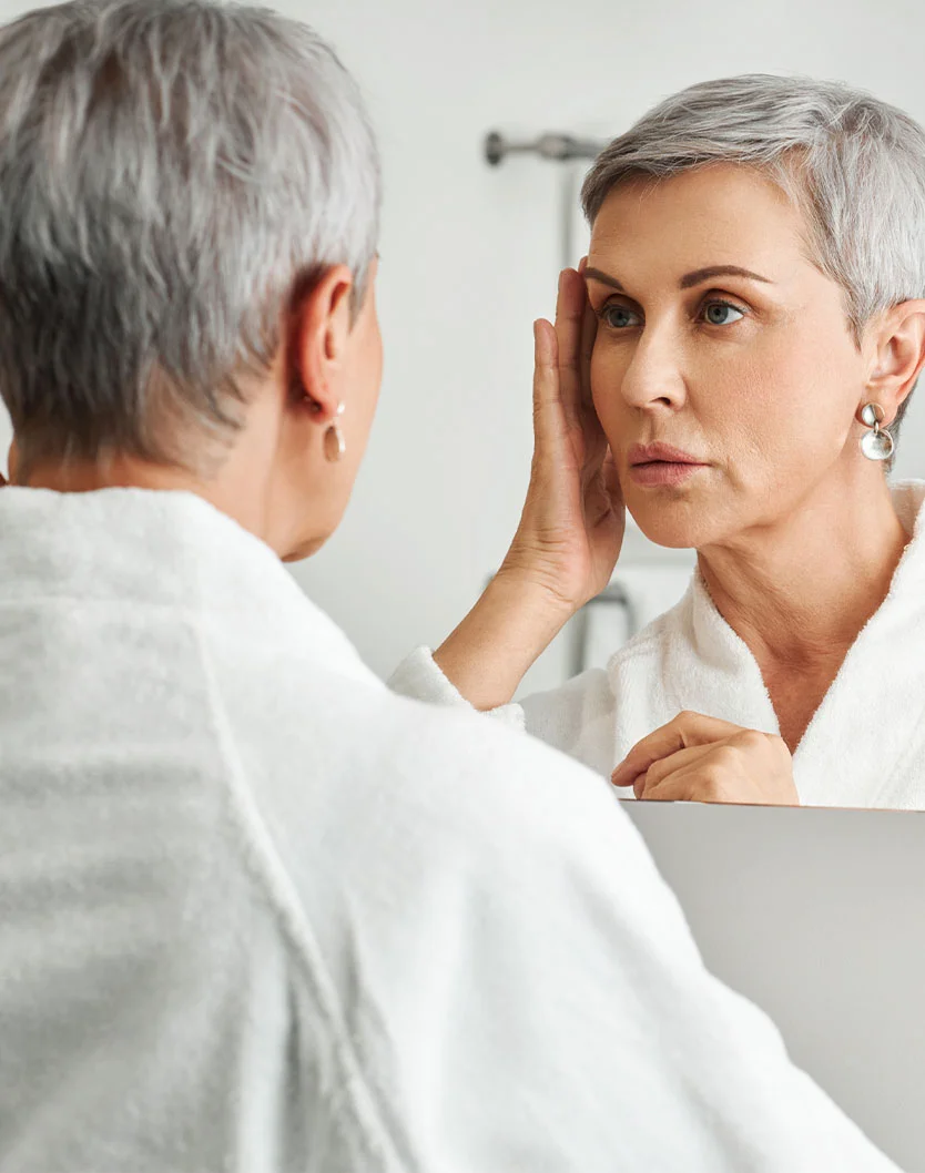 A woman with short grey hair wearing a white robe, looking into a mirror and gently lifting the skin at her temple with her fingers.