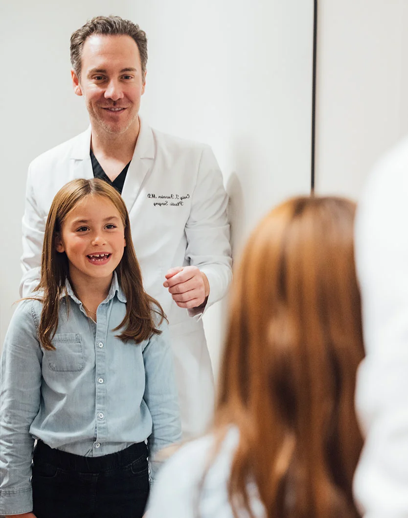 Dr. Fournier in a white lab coat standing behind a young girl in a denim shirt, who is looking into a mirror during a clinical visit.