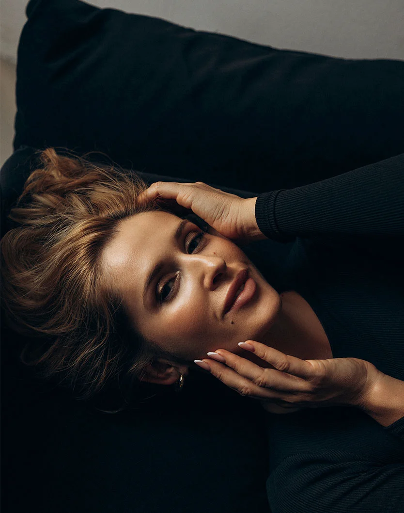 Close-up of a woman lying down on a dark surface, framing her face with her hands to showcase clear, smooth skin texture.
