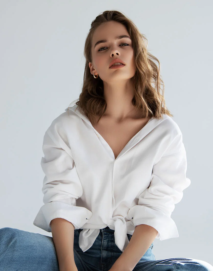 A woman with wavy brown hair wearing an unbuttoned white shirt and jeans, sitting against a white background with a focus on her facial features.