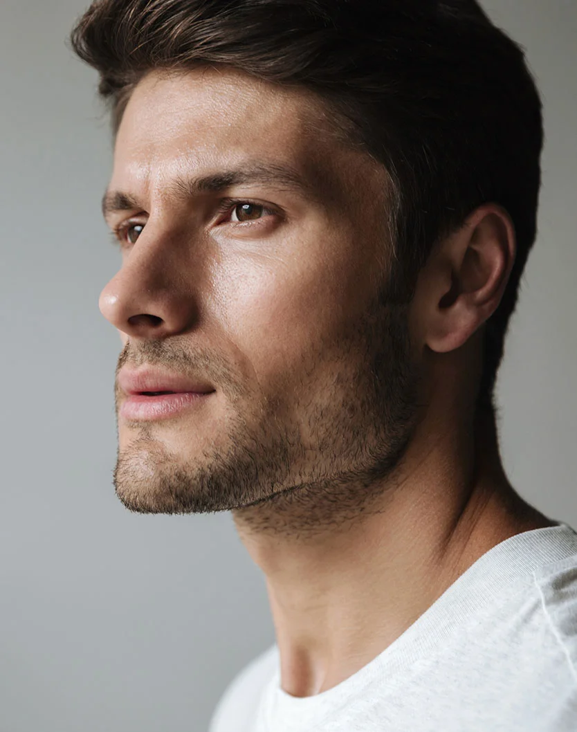 Side profile of a man with short brown hair and light stubble, looking forward against a neutral grey background.