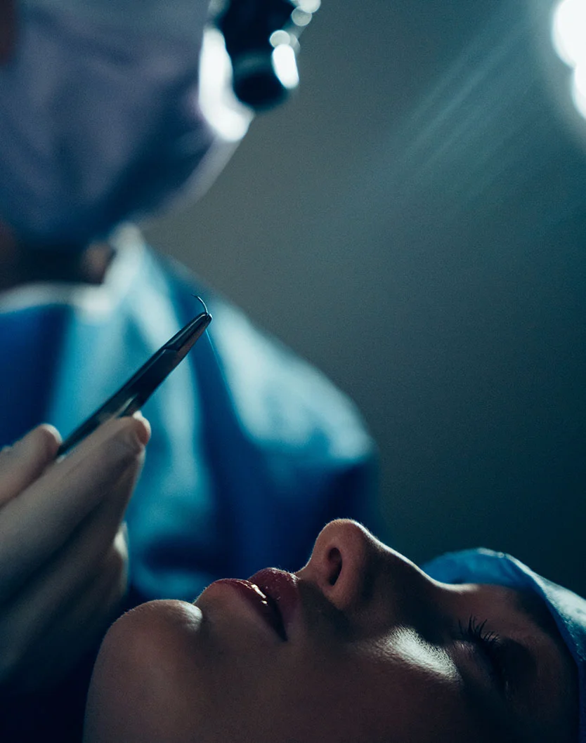 A dramatic, low-light surgical shot of Dr. Fournier in full scrubs and a headlamp, carefully performing a rhinoplasty procedure.