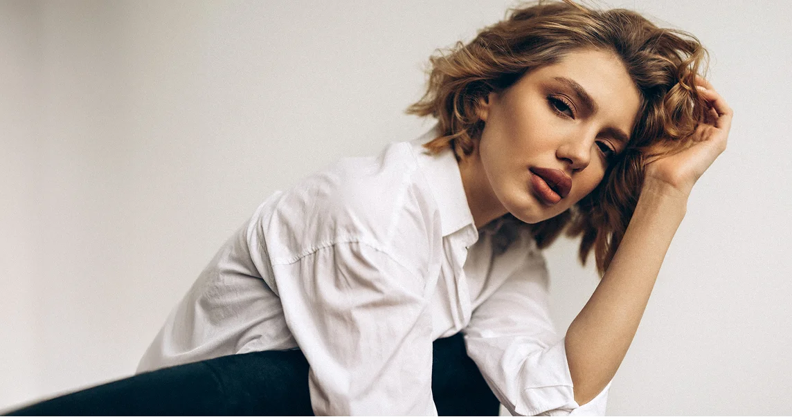 A professional portrait of a woman with wavy blonde hair and soft makeup, wearing an oversized white button-down shirt. She is leaning forward in a thoughtful, artistic pose.
